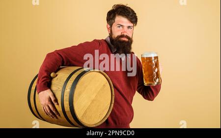 Bearded man with glass of beer and wooden barrel. Tasting fresh beer. Brewer carrying wooden beer keg. Brewery factory. Craft beer. Oktoberfest Stock Photo