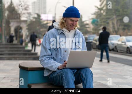 Happy smiling guy student chatting with friends on laptop sitting on bench on city street. Stock Photo
