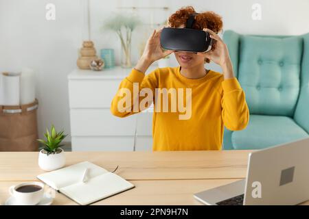 African american young woman wearing using virtual reality metaverse VR glasses headset at home. Girl puts on virtual reality helmet. Technology simulation hi-tech videogame concept Stock Photo