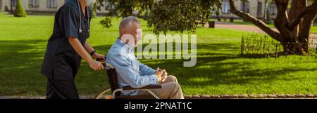Full length shot of caring nurse with senior man in wheelchair on a walk in the park near hospital Stock Photo