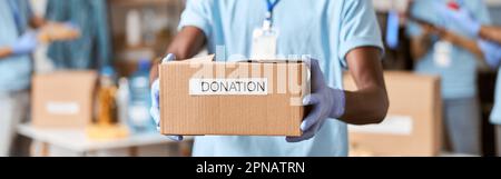 Close up shot of cardboard box for Donation in hands of african american young male volunteer in blue uniform, protective mask and gloves. Team Stock Photo