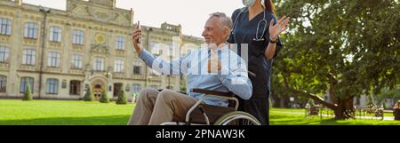 Mature man, recovering patient in wheelchair making a video call using smartphone on a walk with nurse wearing face shield and mask Stock Photo