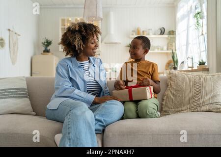 Loving black woman mom congratulate happy small son with birthday give wrapped gift with red ribbon Stock Photo