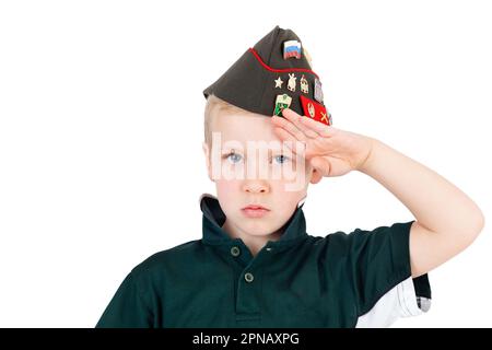 Young caucasian boy saluting playing dressups in green 'army' shirt and russian folding army hat - pilotka - from soviet red army with pins, badges re Stock Photo