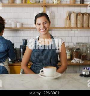 Serving the best coffee in town with a big smile. Portrait of a young waitress standing behind a counter in a cafe. Stock Photo
