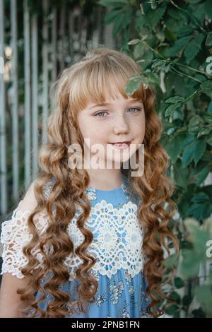 Portrait of a girl with long blonde hair on the street. A 9-11-year-old girl against a background of green plants, smiling and looking at the camera. Stock Photo