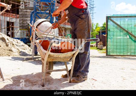 Construction worker is attaching crane hooks to industrial tipping barrow full of red bricks in the background is mortar mixer machine and edifice und Stock Photo