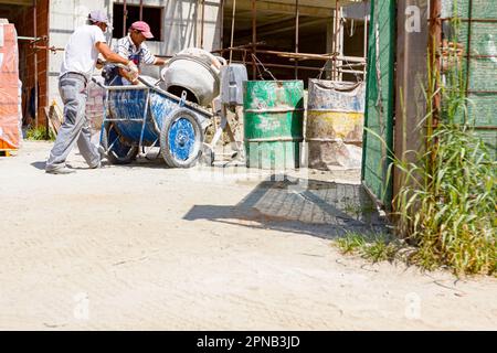 Teamwork, workers are pouring fresh mortar in wheelbarrow from mortar mixer machine. Construction site works Stock Photo