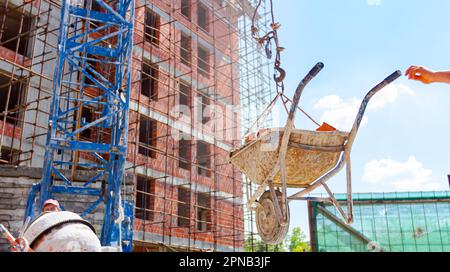 Crane is carrying wheelbarrow full of red bricks, pulled up above building site, keeping balance and direction, in the background is edifice under con Stock Photo