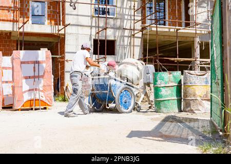 Teamwork, workers are pouring fresh mortar in wheelbarrow from mortar mixer machine. Construction site works Stock Photo