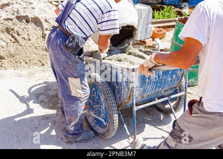 Teamwork, workers are pouring fresh mortar in wheelbarrow from mortar mixer machine. Construction site works Stock Photo