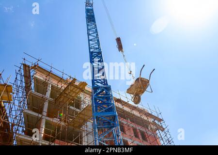 Shot from below of crane that carrying wheelbarrow full of red ceramic blocks, in background is building under construction with scaffolding and blue Stock Photo
