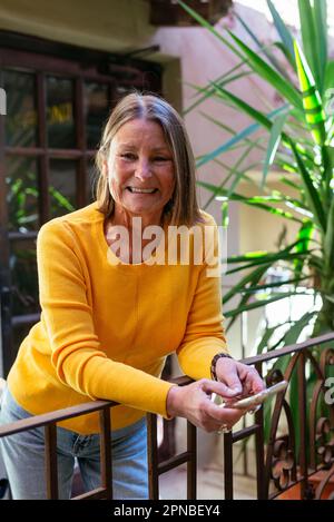 Positive middle aged female with smartphone on hands smiling and looking at camera while leaning on railing of balcony with plants Stock Photo