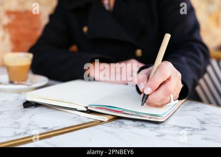 Crop woman in black coat sitting at table with cup of coffee while writing notes on notebook Stock Photo