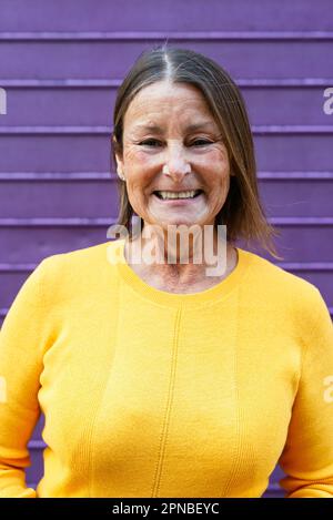 Positive middle aged female in vivid yellow clothes smiling and looking at camera while standing against purple wall Stock Photo