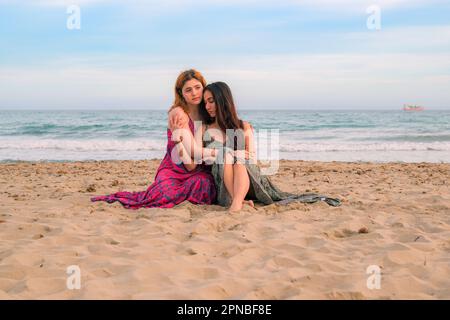Full body of thoughtful young female friends in long dresses sitting on sandy beach near waving sea and hugging while looking away Stock Photo