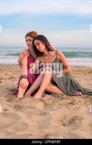 Full body of thoughtful young female friends in long dresses sitting on sandy beach near waving sea and hugging while looking away Stock Photo