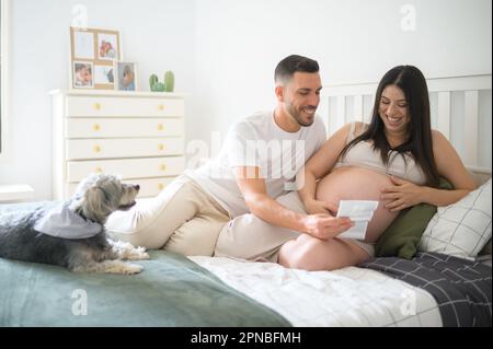 Cheerful young man with pregnant woman smiling and looking at ultrasound photo of baby while sitting on bed near dog in cozy bedroom Stock Photo