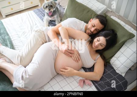 From above of happy young man embracing belly of pregnant smiling woman with closed eyes while lying together on bed near dog in bedroom Stock Photo