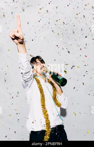 Young unshaven well dressed male with tinsel and bottle of alcoholic drink representing microphone concept closed eyes during party Stock Photo