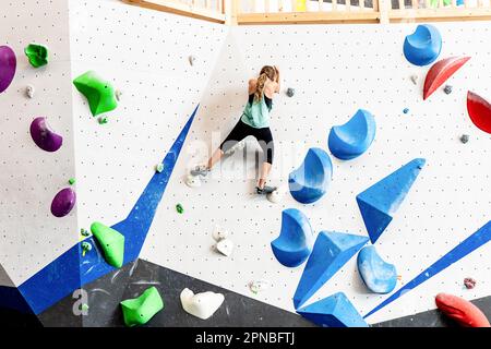 From below back view of unrecognizable female athlete in sportswear and climbing shoes grabbing grips while exercising on climbing wall in bouldering Stock Photo