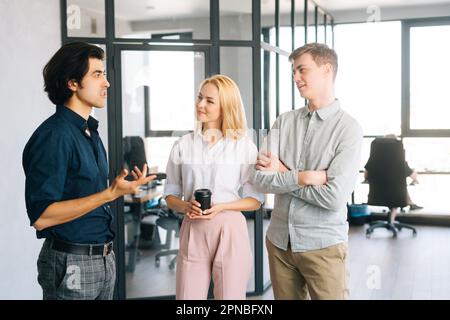 Medium shot of three friendly young business people having fun conversation during coffee break in modern office. Happy coworkers enjoy pleasant Stock Photo