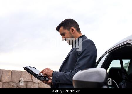 Side view of happy bearded male entrepreneur in classy suit leaning on car while browsing tablet and looking at screen against blue sky Stock Photo