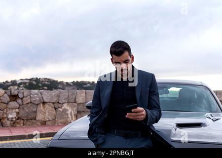 Young male worker in formal suit browsing mobile phone and looking at screen while leaning on car on street Stock Photo