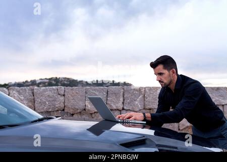 Side view of young bearded male entrepreneur in formal clothes working with laptop placed on car hood Stock Photo