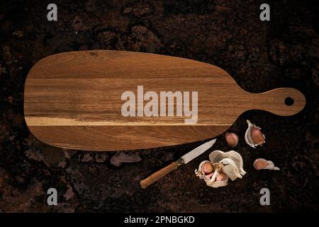 Top view of wooden oval shaped cutting board placed near knife and garlic on rough surface Stock Photo