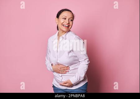 Delightful pregnant woman putting hands on her belly, feeling first baby touches, smiling expressing positive emotions Stock Photo