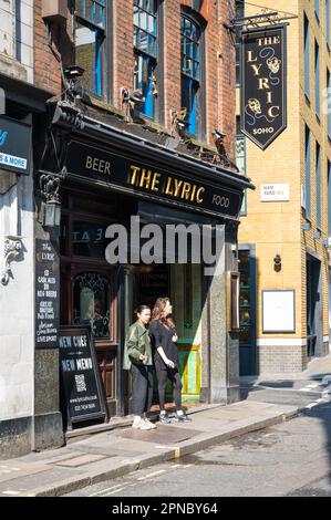 Two young women stand smoking cigarettes outside The Lyric pub. Great Windmill Street, Soho, London, England, UK Stock Photo