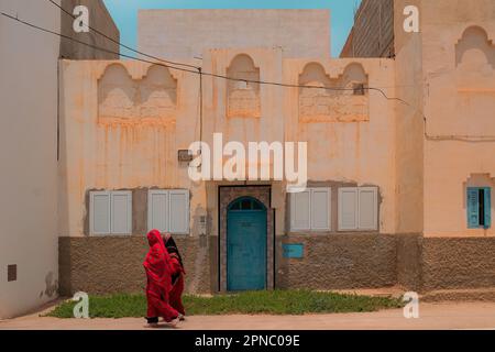 Two local Muslim women covered in red Abayas walk past an old house on a sidewalk in Sidi Ifni, Morocco during Ramadan. Traditional religious clothing. Stock Photo