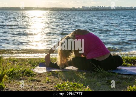 A woman in sports clothes, sitting in the lotus position and bending her hand to her leg on a rug on the lake shore. Meditation. Sports in old age. Yo Stock Photo