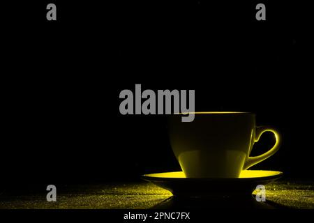 A mug with a warm drink illuminated by yellow light on black background, silhouette, copy space, creative. Morning coffee Stock Photo