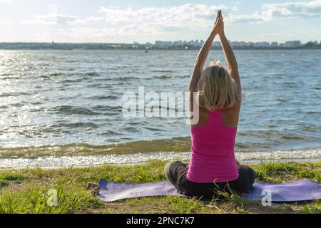 An elderly woman sitting in the lotus position and raising her hands up and looking at the lake, a view from the back. Meditation. Sports in old age. Stock Photo