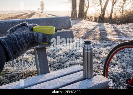 Hand in a mitten holds a mug with a hot drink, against the background of a thermos, which stands on a bench, on a frosty morning.  Stock Photo