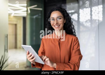 Portrait of successful hispanic woman inside office at workplace, businesswoman smiling and looking at camera, female worker holding tablet computer. Stock Photo