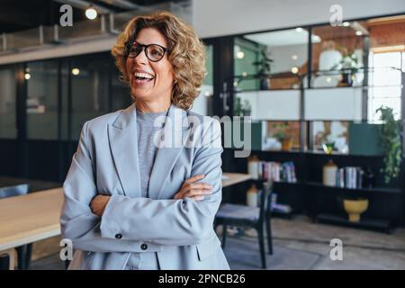Mature business woman smiling and standing with crossed arms. Professional business woman wearing a suit in an office. Stock Photo