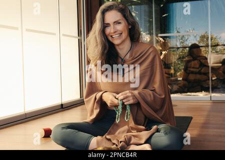 Mature woman smiling at the camera while holding a beads necklace. Happy senior woman using mala beads to meditate at home. Cheerful woman practicing Stock Photo