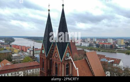 18 April 2023, Brandenburg, Frankfurt (Oder): Two employees from the company Bittner Glocken- und Turmuhren and a technician from the lifting platform mount the gilded hands and numerals on the southern tower clock of the Friedenskirche at a height of 38 meters (aerial view with a drone). On the same day, two employees of the Bittner company had remounted the restored and newly gilded hands and numerals of the church tower clock, which dates back to about 1900, on the clock face. The clockwork had already been restored by the company. The restoration project around the tower clock arose in 201 Stock Photo