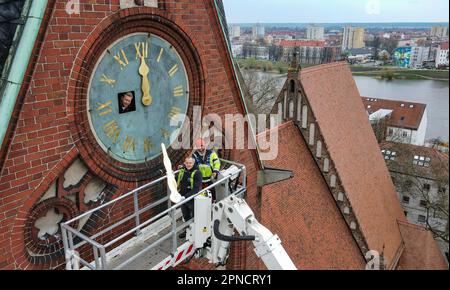 18 April 2023, Brandenburg, Frankfurt (Oder): Two employees from the company Bittner Glocken- und Turmuhren and a technician from the lifting platform mount the gilded hands and numerals on the southern tower clock of the Friedenskirche at a height of 38 meters (aerial view with a drone). On the same day, two employees of the Bittner company had remounted the restored and newly gilded hands and numerals of the church tower clock, which dates back to about 1900, on the clock face. The clockwork had already been restored by the company. The restoration project around the tower clock arose in 201 Stock Photo