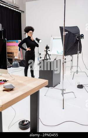 pensive african american content maker standing with hand on hip near laptop and softbox reflector in photo studio,stock image Stock Photo