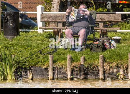 Hickling Basin Lincolnshire 18th April  2023 :  Fisherman reading the newspaper clear blue sky light wind enjoying the relaxing sounds and views of mother nature  Chinook  helicopter  overhead  :Clifford Norton  Alamy Live News Stock Photo
