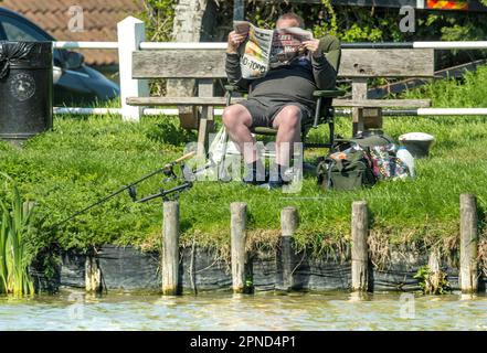 Hickling Basin Lincolnshire 18th April  2023 :  Fisherman reading the newspaper clear blue sky light wind enjoying the relaxing sounds and views of mother nature  Chinook  helicopter  overhead  :Clifford Norton  Alamy Live News Stock Photo