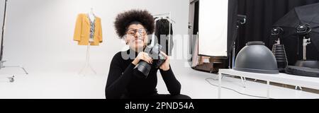 joyful african american photographer in eyeglasses smiling with closed eyes while sitting in photo studio, banner,stock image Stock Photo