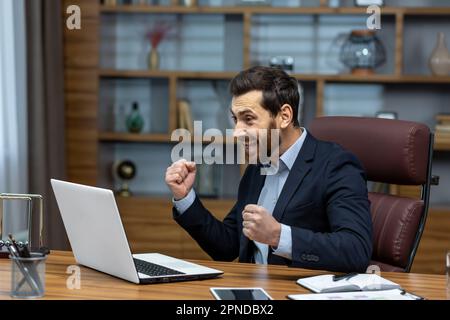 Successful businessman in home office celebrating victory and success, mature adult male boss satisfied with achievement results holding hands up triumph gesture financier happy using laptop at work Stock Photo