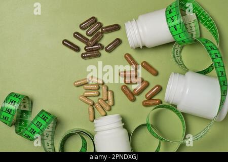 Various capsules with vitamins or dietary supplements spill out of three white plastic jars wrapped with a measure tape on a green background Stock Photo