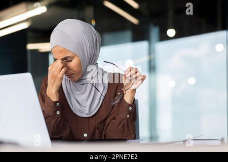Tired young Muslim female teacher in hijab sits at desk in front of laptop. She holds glasses in her hand, closed her eyes, rubs her eyes with her hand, rests, takes a break in online learning. Stock Photo