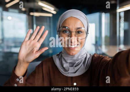 Close-up photo. A young female blogger in a hijab talks to the camera, holds a phone in her hand. Films a blog, a podcast. He greets the camera with his hand, smiles. Stock Photo